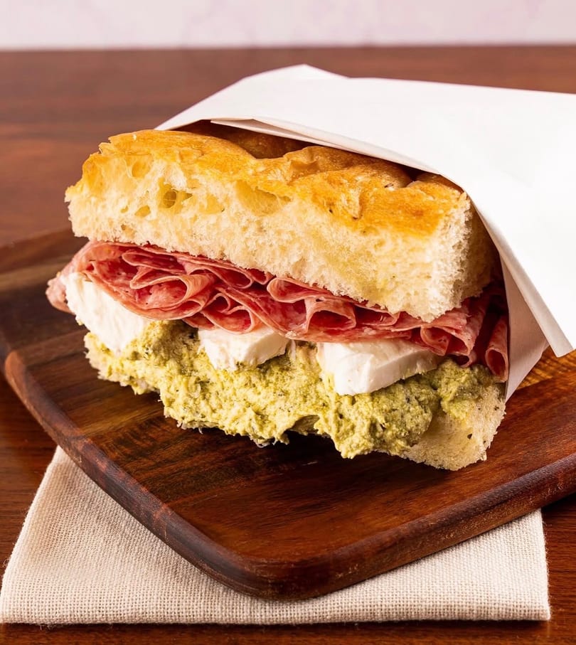 Focaccia sandwich with salami, mozzarella, and pesto on a wooden board.