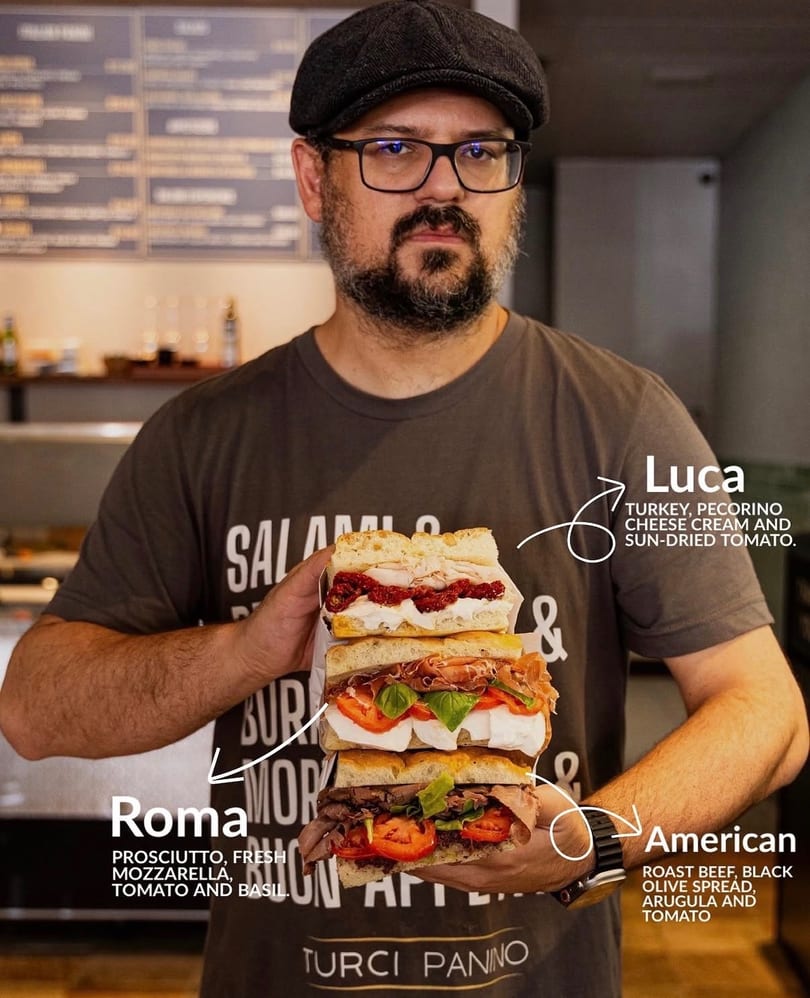 Man in a flat cap and glasses holding a stack of three gourmet sandwiches from Turci Panino.