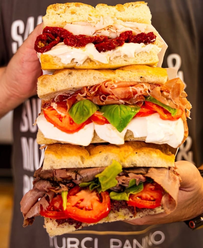 A person holding a towering stack of three focaccia sandwiches with various fillings.