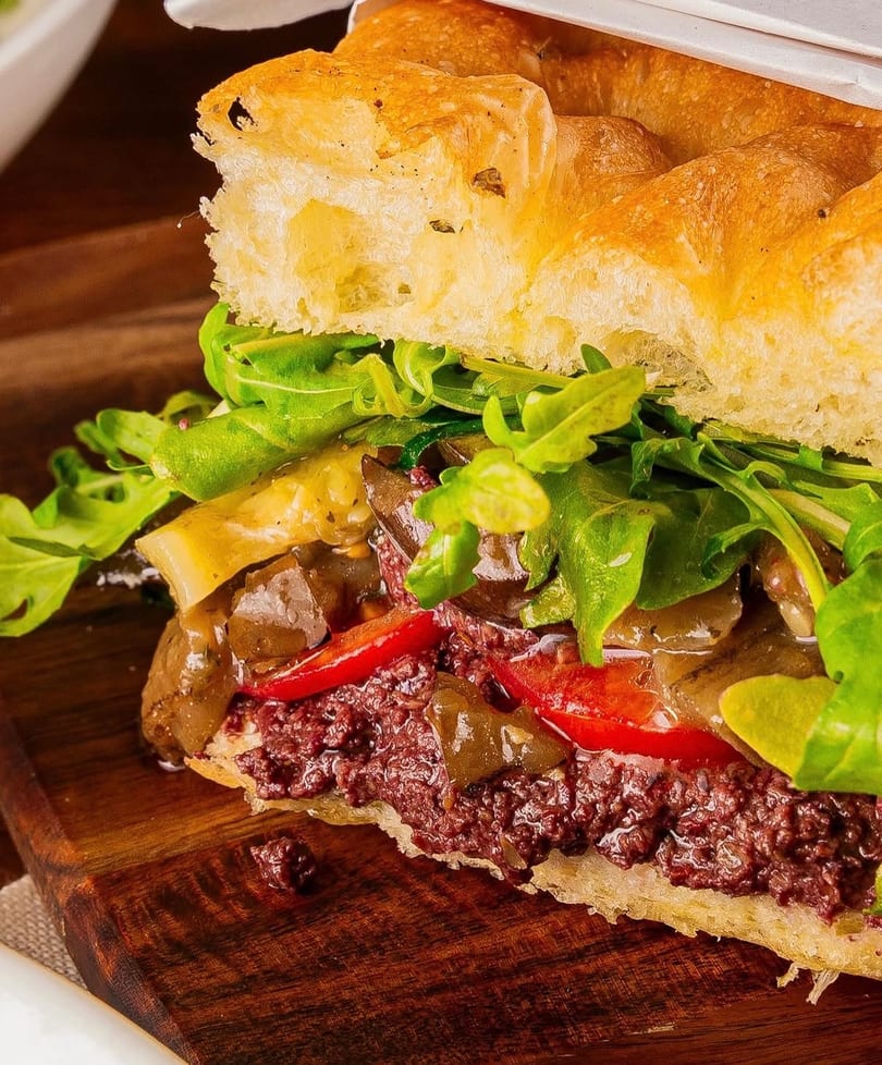 Close-up of a focaccia sandwich filled with olive tapenade, roasted peppers, and arugula on a wooden board.
